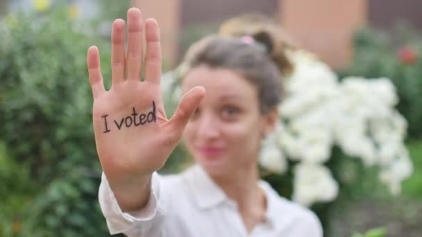 Ao ar livre retrato feminino de jovem mulher adulta em camisa branca mostrando sua mão com slogan Eu votei no fundo flores, conceito dia eleições — Vídeo de Stock