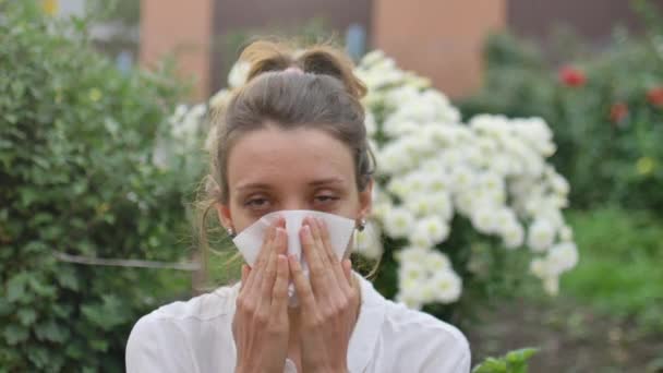 Girl sneezes into a napkin, because she is allergic to flowering during spring time on white chrysanthemums on background, hypersensitivity concept — стоковое видео