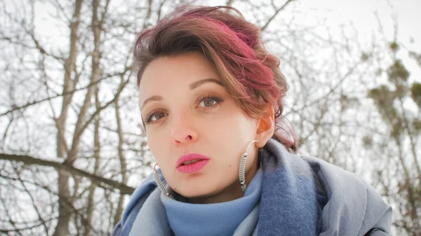 Привлекательная молодая женщина с идеальной кожей и макияжем с темно-розовыми волосами обладает на фоне зимнего парка носить синий шарф и серебряные серьги — стоковое фото