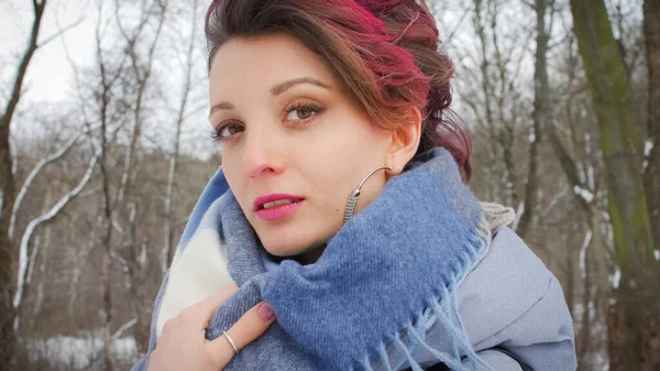 Jovem atraente com pele perfeita e maquiagem com cabelo rosa escuro está possuindo no fundo do parque de inverno vestindo cachecol azul e brincos de prata — Fotografia de Stock