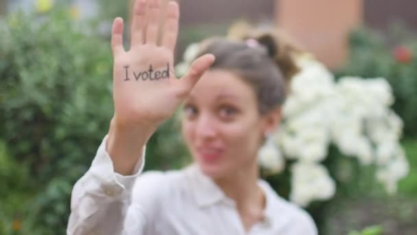 На відкритому повітрі жіночий портрет молодої дорослої жінки в білій сорочці, що показує її руку з гаслом Я голосував на квітковому фоні, концепція дня виборів — стокове відео