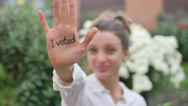Utomhus kvinnligt porträtt av ung vuxen kvinna i vit skjorta visar sin hand med slogan Jag röstade på blommor bakgrund, val dag koncept — Stockvideo