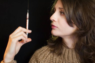 Aşı konsepti. Kafkasyalı bir kızın elinde şırınga tutan bir kadın portresi. Stüdyodaki siyah arka planda ona bakıyor..