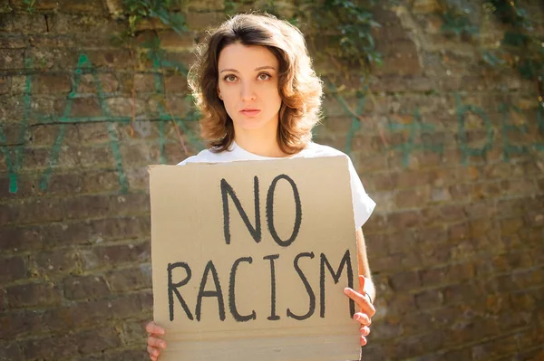 Upset young protesting woman in white shirt holds protest sign broadsheet placard with slogan No racism for public demonstration on stone wall background