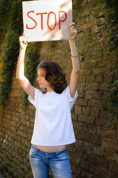 Young protesting woman in white shirt and jeans holds protest sign broadsheet placard with slogan Stop for public demonstration on wall background. — Stock Photo, Image