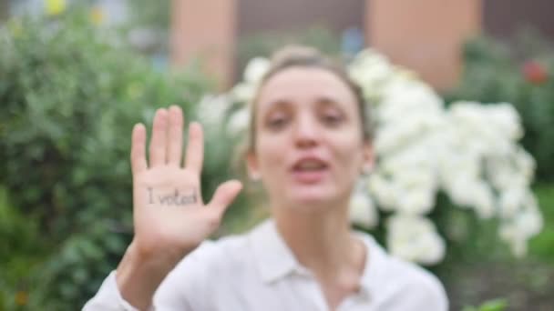Ao ar livre retrato feminino de jovem mulher adulta em camisa branca mostrando sua mão com slogan Eu votei no fundo flores, conceito dia eleições — Vídeo de Stock