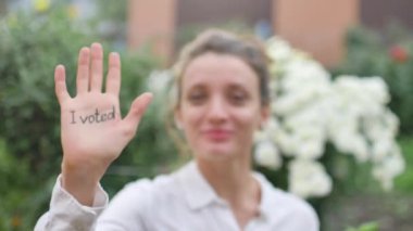 Açık havada beyaz tişörtlü, elini gösteren kadın portresi. Çiçeklere arka planda oy verdim. Seçim günü konsepti.