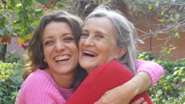Yaşlı anne, yetişkin kızıyla birlikte bahçedeki kameraya bakıyor ve güneşli bir günde birbirlerine sarılıyorlar.