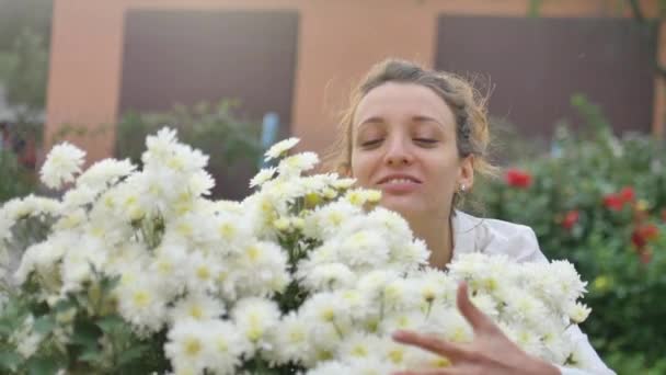 Досить молода дівчина стоїть біля куща білих хризантем в парку під час весни, гіперчутливості, астми або алергії концепції — стокове відео
