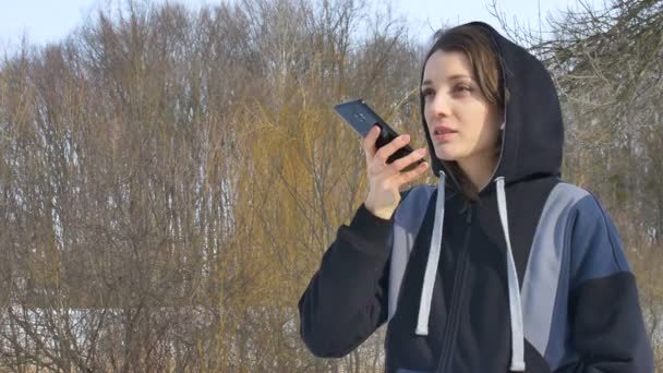 Junge Fitnesstrainerin in dunkler Sportbekleidung schickt Audio-Sprachnachricht, die auf dem Smartphone im Freien etwas erklärt, in ein neues soziales Netzwerk — Stockvideo