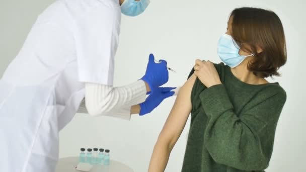 Kvinnlig läkare eller sjuksköterska ger en spruta eller vaccin till en patient skuldra av ung kvinna i grön tröja och skyddande ansiktsmask. Vaccination och förebyggande av influensa eller viruspandemi. — Stockvideo
