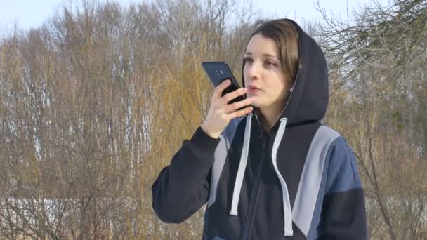 Joven entrenador de fitness en ropa deportiva oscura está enviando un mensaje de voz de audio explicando algo en el teléfono inteligente al aire libre a una nueva red social — Vídeos de Stock