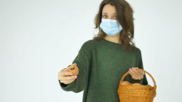 Junge Frau in grünem Pullover und Schutzmaske zeigt handgezeichnete Eier mit anthropomorphem Smiley-Gesicht aus Strohkorb, Ostervorbereitung, Urlaubsstimmung während der Pandemiezeit — Stockvideo