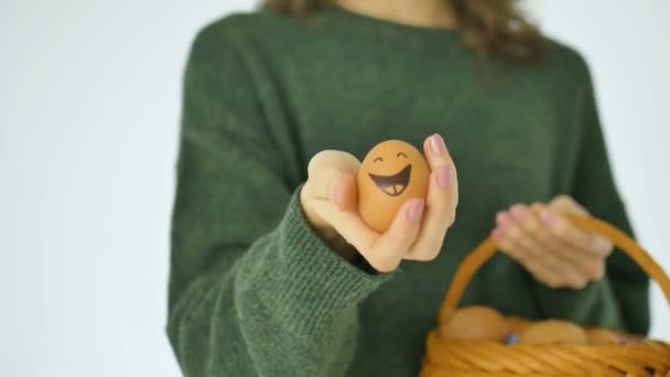 緑のセーターの若い女性は、藁バスケット、イースター準備、休日の気分の概念から人形の笑顔の顔を持つ手描きの卵を示しています — ストック動画