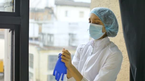 파란색 장갑을 끼고 보호 복을 입은 여성 간호사가 코로나 바이러스 대유행 이 끝 나기를 기다리면서 창문 근처에서 마스크를 벗고 있다 — 비디오