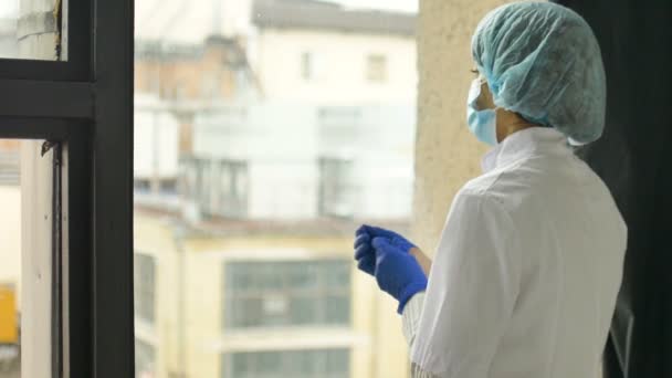 Portrett av en ulykkelig, ung lege som har hodepine ved et vindu. Viktig jobb og selvisolering under coronavirus pandemi. Håp om medisin. – stockvideo