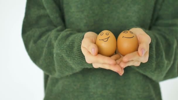 Νεαρή γυναίκα σε πράσινο πουλόβερ δείχνει τα αυγά με ζωγραφισμένο στο χέρι πρόσωπο με ένα χαμόγελο από ψάθινο καλάθι, προετοιμασία Πάσχα, τις έννοιες διάθεση διακοπών — Αρχείο Βίντεο