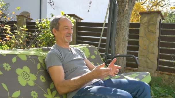 Hombre mayor está sentado en un columpio al aire libre durante el día soleado de verano en el patio de recreo en el jardín y pensando en algo — Vídeos de Stock