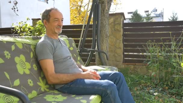 Hombre mayor está sentado en un columpio al aire libre durante el día soleado de verano en el patio de recreo en el jardín y pensando en algo — Vídeos de Stock