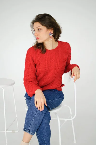 Studioporträt einer jungen Frau in rotem Strickpullover und Ohrring, die auf einem Stuhl auf weißem Hintergrund sitzt — Stockfoto