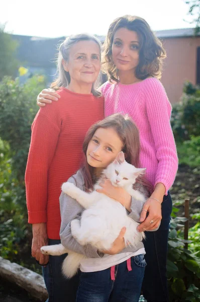 Madre mayor con el pelo gris con su hija adulta y su nieta pequeña con gato blanco están mirando a la cámara en el jardín y abrazándose durante el día soleado al aire libre —  Fotos de Stock