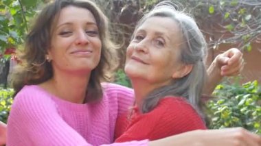 Yaşlı anne ve yetişkin kızı bahçede güneşli bir günde birbirlerine sarılıyorlar.