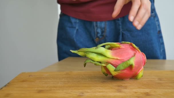 Le mani femminili stanno tagliando un frutto di drago o pitaya con pelle rosa e polpa bianca con semi neri sul tagliere di legno sul tavolo. Frutti esotici, concetto di alimentazione sana — Video Stock