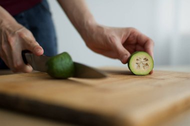 Dişi eller kesilmiş bir tahta üzerinde taze yeşil bir feijoa meyvesi kesiyor. Egzotik meyveler, sağlıklı beslenme konsepti