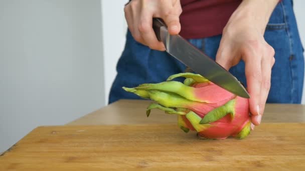 Le mani femminili stanno tagliando un frutto di drago o pitaya con pelle rosa e polpa bianca con semi neri sul tagliere di legno sul tavolo. Frutti esotici, concetto di alimentazione sana — Video Stock