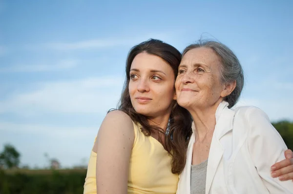 Madre mayor con el pelo gris con su hija adulta mirando a la cámara en el jardín y abrazándose durante el día soleado al aire libre —  Fotos de Stock