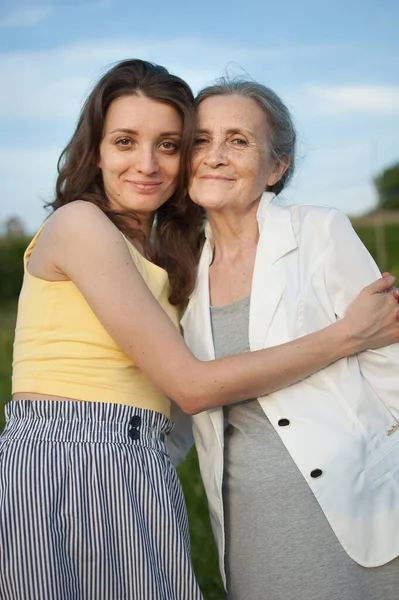 Madre mayor con el pelo gris con su hija adulta mirando a la cámara en el jardín y abrazándose durante el día soleado al aire libre —  Fotos de Stock