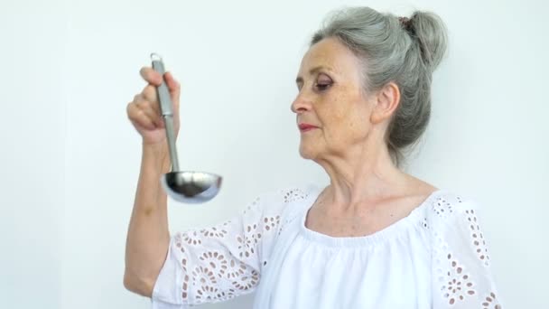 Femme âgée émotionnelle aux cheveux argentés tient une louche ou une cuillère en métal sur fond blanc, retraite heureuse, concepts de la fête des mères. — Video