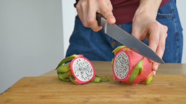 Las manos femeninas están cortando una fruta de dragón o pitaya con piel rosa y pulpa blanca con semillas negras sobre tabla de cortar de madera en la mesa. Frutas exóticas, concepto de alimentación saludable — Vídeos de Stock