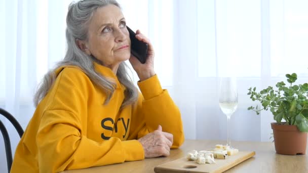 Belle vieille grand-mère avec les cheveux gris et le visage avec des rides utilise smartphone, parler avec quelqu'un et assis à la table à la maison sur fond de fenêtre, fête des mères, retraite heureuse — Video