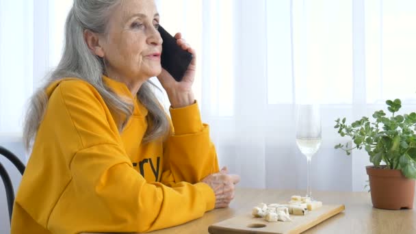 Belle vieille grand-mère avec les cheveux gris et le visage avec des rides utilise smartphone, parler avec quelqu'un et assis à la table à la maison sur fond de fenêtre, fête des mères, retraite heureuse — Video