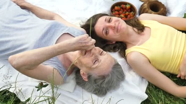 Красивая старая бабушка с седыми волосами и лицом с морщинами лежит на белом ковре со своей взрослой дочерью на зеленой траве во время пикника на открытом воздухе, День матери, счастливого выхода на пенсию — стоковое видео