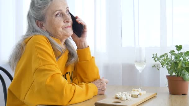 Belle vieille grand-mère avec les cheveux gris et le visage avec des rides utilise smartphone, parler avec quelqu'un et assis à la table à la maison sur fond de fenêtre, fête des mères, retraite heureuse — Video