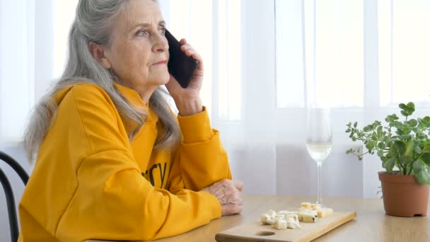 Belle vieille grand-mère avec les cheveux gris et le visage avec des rides utilise smartphone, parler avec quelqu'un et assis à la table à la maison sur fond de fenêtre, fête des mères, retraite heureuse — Video