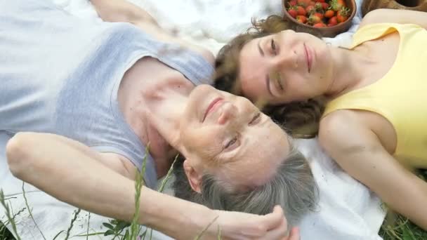 Όμορφη γιαγιά με γκρίζα μαλλιά και το πρόσωπο με ρυτίδες βρίσκεται σε λευκό χαλί με την ενήλικη κόρη της στο πράσινο γρασίδι κατά τη διάρκεια πικ-νικ σε εξωτερικούς χώρους, ημέρα μητέρες, ευτυχισμένη συνταξιοδότηση — Αρχείο Βίντεο