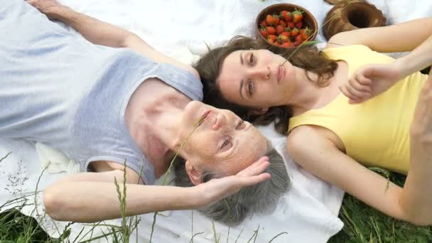 Beautiful old grandmother with grey hair and face with wrinkles is lying on white carpet with her adult daughter on green grass during picnic outdoors, mothers day, happy retirement — Stock Video