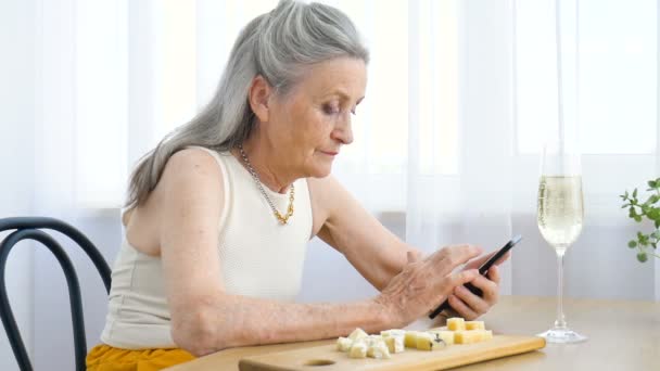 灰色の髪としわの顔を持つ美しい祖母は、スマートフォンを使用しています,誰かと話をし、ウィンドウの背景に自宅でテーブルに座って,母の日,幸せな退職 — ストック動画