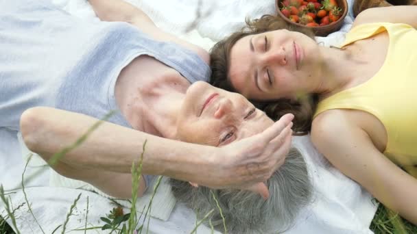 Όμορφη γιαγιά με γκρίζα μαλλιά και το πρόσωπο με ρυτίδες βρίσκεται σε λευκό χαλί με την ενήλικη κόρη της στο πράσινο γρασίδι κατά τη διάρκεια πικ-νικ σε εξωτερικούς χώρους, ημέρα μητέρες, ευτυχισμένη συνταξιοδότηση — Αρχείο Βίντεο