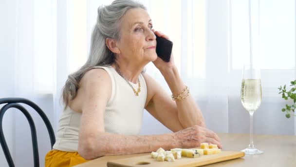 Belle vieille grand-mère avec les cheveux gris et le visage avec des rides utilise smartphone, parler avec quelqu'un et assis à la table à la maison sur fond de fenêtre, fête des mères, retraite heureuse — Video