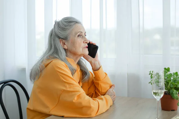 Beautiful old grandmother with grey hair and face with wrinkles is using smartphone, talking with someone and sitting at the table at home on window background, mothers day, happy retirement