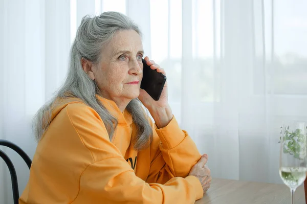 Beautiful old grandmother with grey hair and face with wrinkles is using smartphone, talking with someone and sitting at the table at home on window background, mothers day, happy retirement — Stock Photo, Image