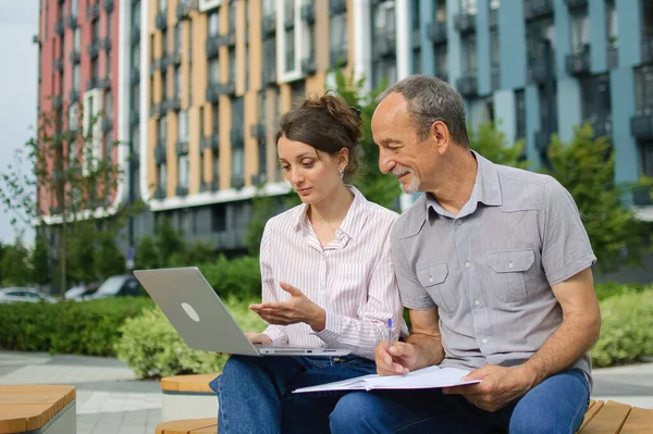 Attractive young woman and senior man are using laptop sitting on the bench in modern residential complex. Casual meeting outside office. A daughter is teaching technology for her father
