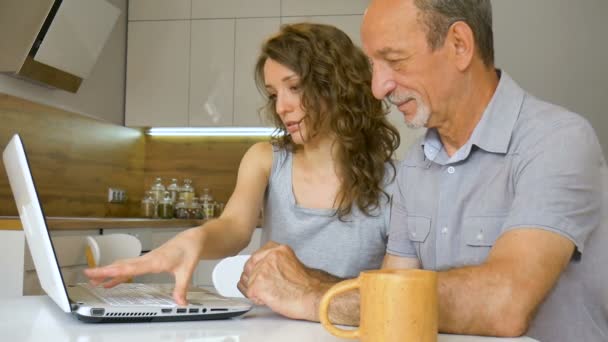 Attrayant jeune fille et père aîné utilisent ordinateur portable assis dans la cuisine moderne dans l'appartement. Femme enseigne la technologie pour son père à la maison, concept de retraite heureuse — Video