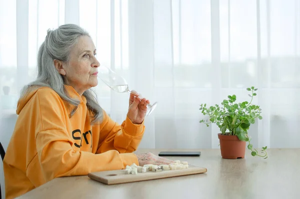 Hermosa abuela de edad con el pelo gris y la cara con arrugas sentado a la mesa en casa en el fondo de la ventana con una copa de champán, día de las madres, feliz jubilación —  Fotos de Stock