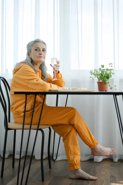 Hermosa abuela de edad con el pelo gris y la cara con arrugas sentado a la mesa en casa en el fondo de la ventana con una copa de champán, día de las madres, feliz jubilación —  Fotos de Stock