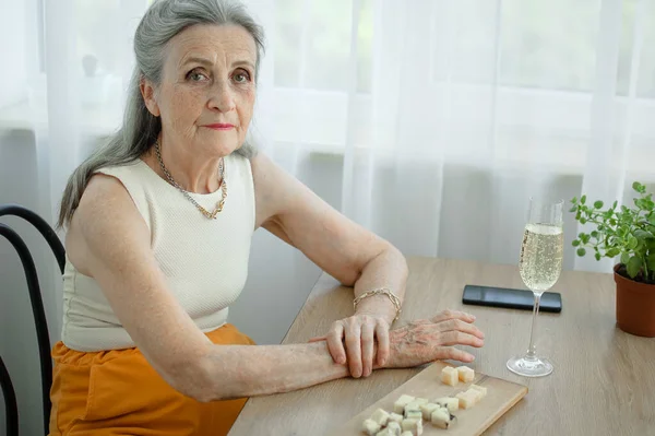 Beautiful old grandmother with grey hair and face with wrinkles sitting at the table at home on window background with glass of champagne, mothers day, happy retirement — Stock Photo, Image